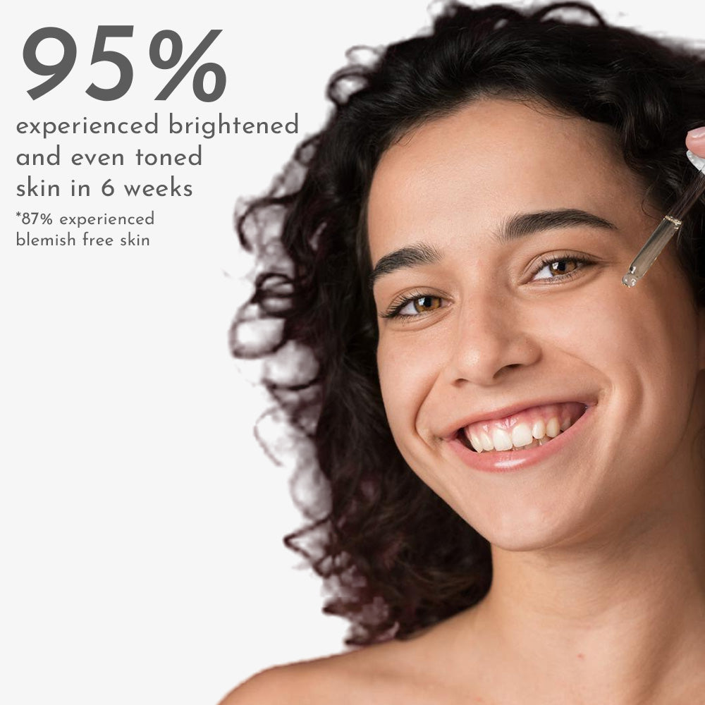 A woman with curly dark hair smiles as she applies best vitamin c serum from a dropper bottle to her cheek.