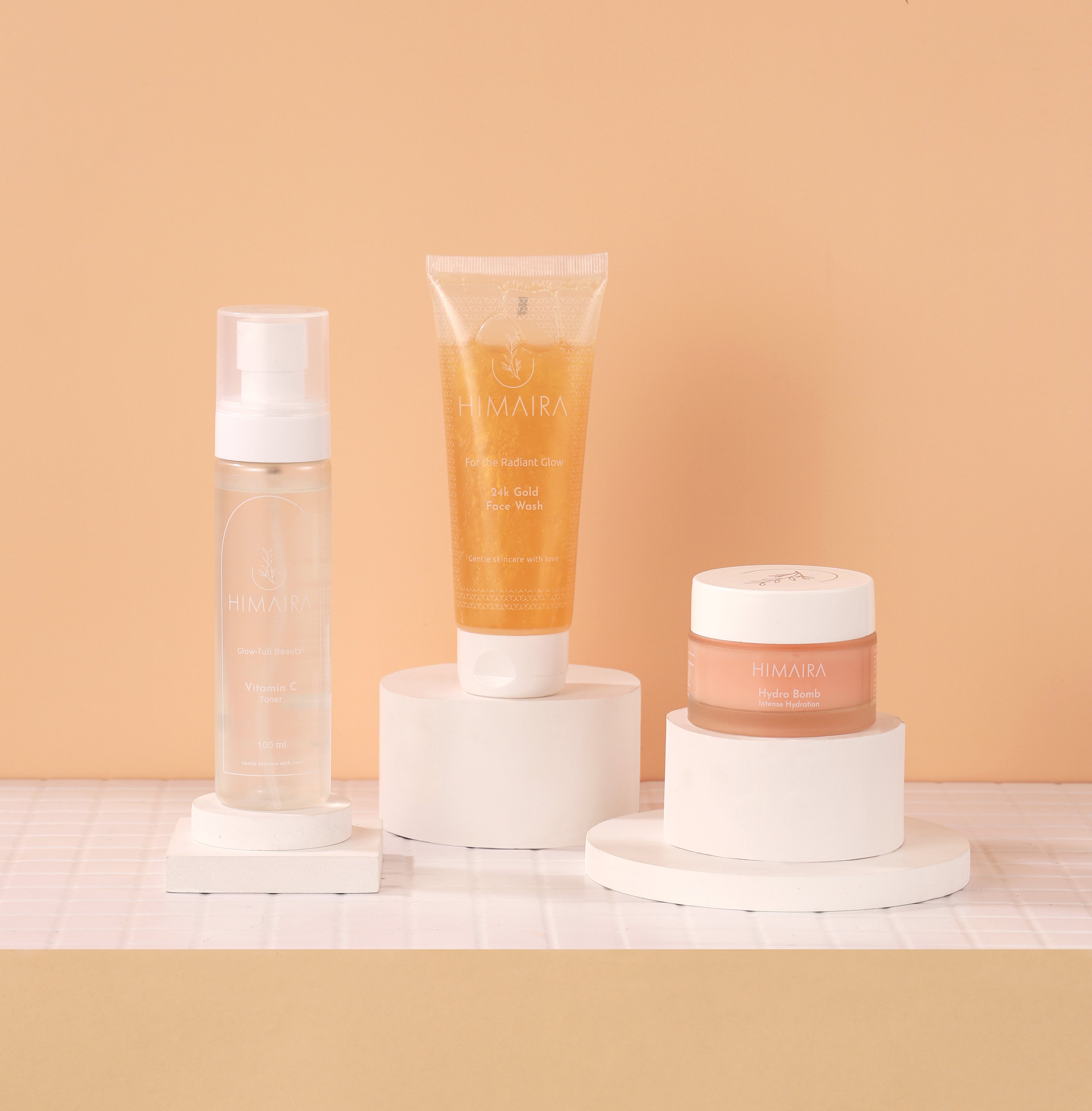 Product lineup with yellow color Vitamin C Face Wash tube, clear Vitamin C Toner and peach color jar of Hydro Bomb moisturiser displayed on white blocks and ring.